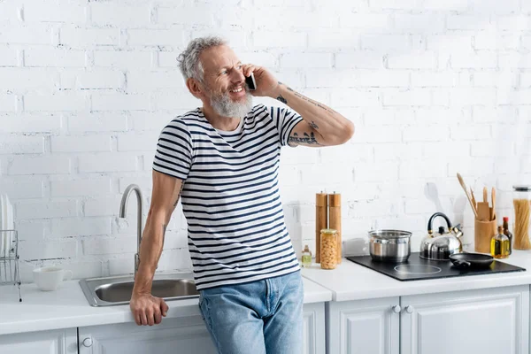 Glücklicher älterer Mann spricht auf Smartphone neben Arbeitsplatte in Küche — Stockfoto