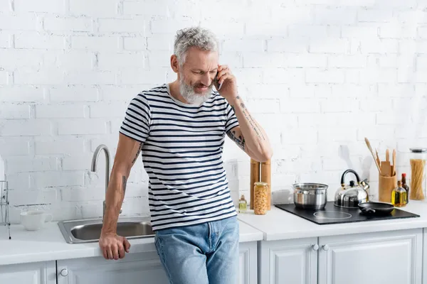 Mature man talking on cellphone in kitchen. Translation: 