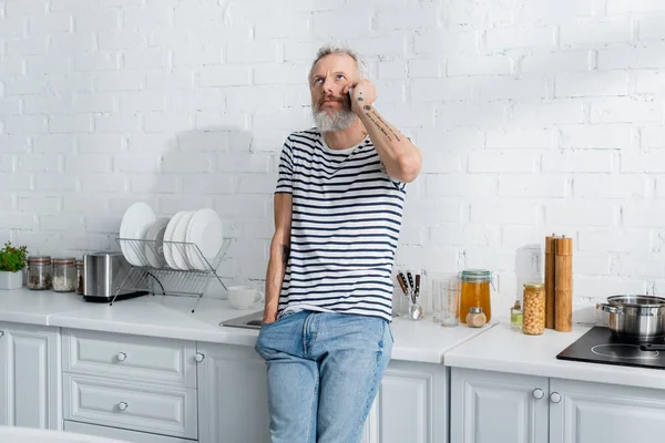 Pensive mature tattooed man talking on smartphone near worktop in kitchen — Stock Photo