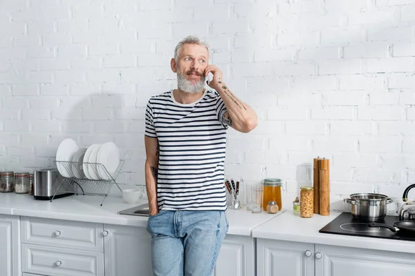 Middle aged man in striped t-shirt talking on mobile phone in kitchen — Stock Photo