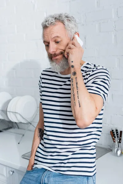 Displeased mature man talking on smartphone near worktop in kitchen — Stock Photo