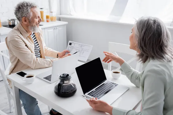 Mujer madura señalando facturas cerca de marido con portátil y café en la cocina - foto de stock