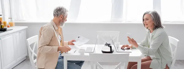 Pareja madura cuenta facturas cerca de café y dispositivos en la cocina, pancarta - foto de stock