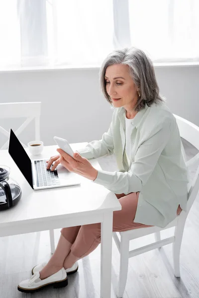 Mature woman using laptop and smartphone near coffee at home — Stock Photo