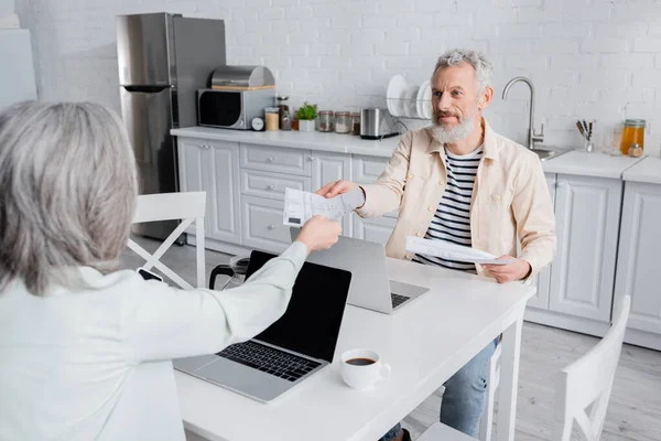 Mature man taking bills to wife near cup of coffee and laptops at home — Stock Photo