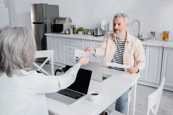 Homme mûr prenant des factures à la femme près de tasse de café et ordinateurs portables à la maison — Photo de stock