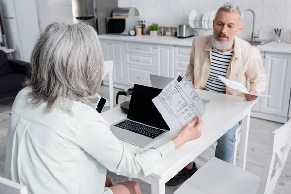 Mature woman holding bills near devices and husband in kitchen — Stock Photo