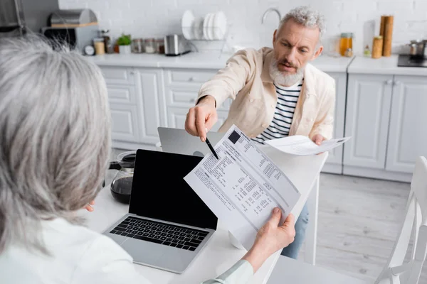 Uomo maturo indicando fatture vicino moglie e computer portatili a casa — Foto stock