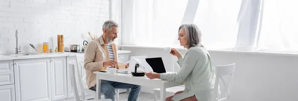 Mujer madura sosteniendo taza de café y facturas cerca del marido con el ordenador portátil en la cocina, pancarta - foto de stock