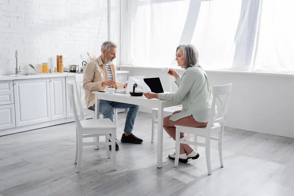 Ältere Frau schaut sich Rechnungen in der Nähe von Kaffee, Laptops und Ehemann in der Küche an — Stockfoto