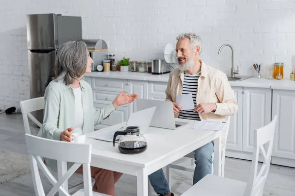 Sorrindo homem segurando contas perto da esposa e laptops na cozinha — Fotografia de Stock