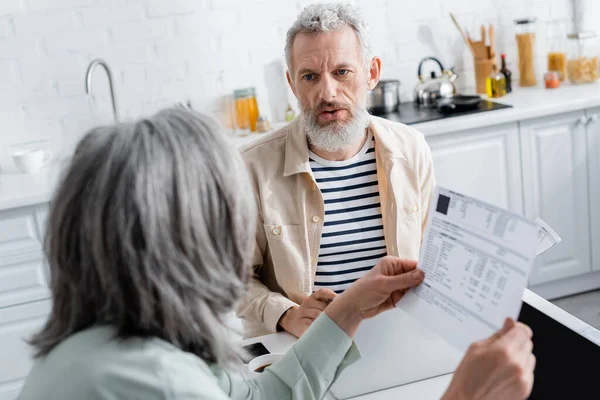 Mature man talking to wife with bills near laptops and smartphone in kitchen — Stock Photo