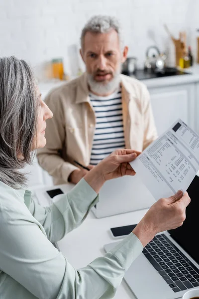 Mujer madura sosteniendo papel con cuentas cerca de dispositivos y marido borroso en la cocina - foto de stock