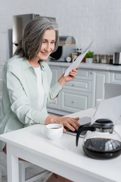 Mujer sonriente sosteniendo facturas y el uso de ordenador portátil cerca de café en la cocina - foto de stock