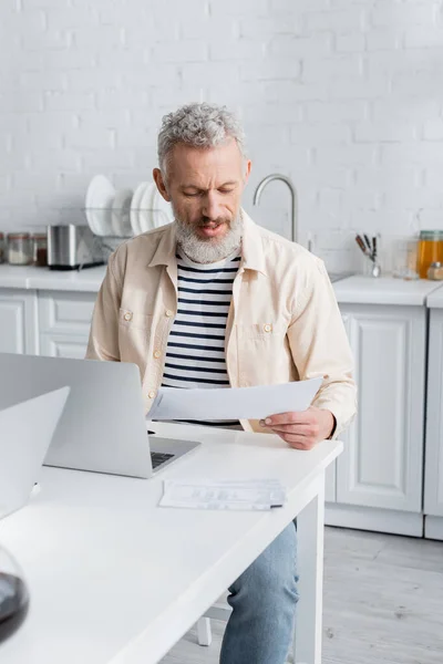 Reifer Mann schaut sich in der Küche zu Hause Papiere in der Nähe von Laptop an — Stockfoto
