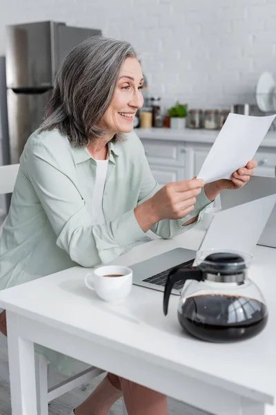 Mujer alegre sosteniendo papel cerca de café y portátil en casa - foto de stock