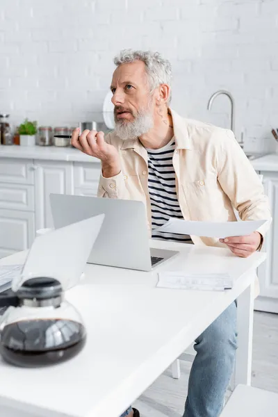 Pensativo hombre maduro sosteniendo papeles cerca de computadoras portátiles y cafetera en casa - foto de stock