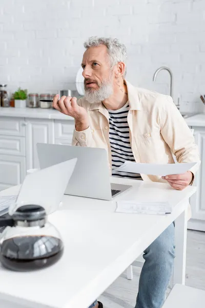 Uomo maturo pensieroso che tiene carte vicino a computer portatili e caffettiera a casa — Foto stock