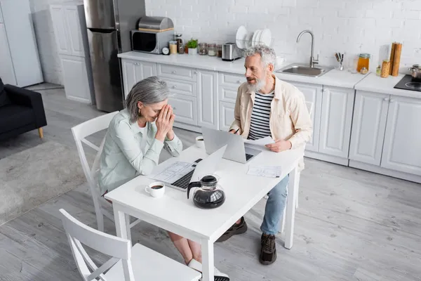 Sorridente coppia matura seduta vicino a bollette, computer portatili e caffè in cucina — Foto stock