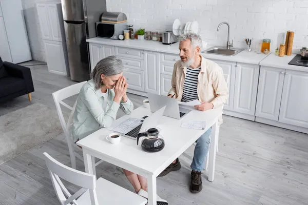 Pareja madura sonriente sentada cerca de facturas, laptops y café en la cocina - foto de stock