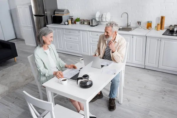 Alegre pareja madura mirándose cerca de facturas, café y computadoras portátiles en casa - foto de stock