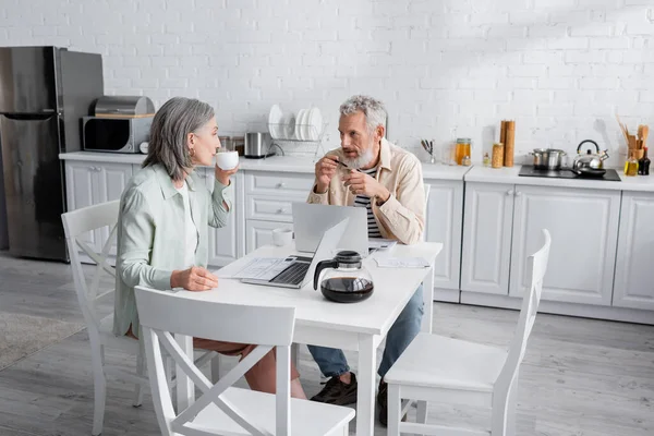 Homem de meia idade olhando para a esposa com xícara de café perto de contas e laptops na cozinha — Fotografia de Stock