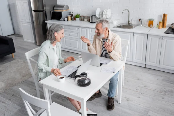 Happy mature woman looking at husband near bills, coffee and laptops in kitchen — Stock Photo