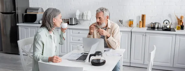 Hombre maduro hablando con su esposa con café cerca de computadoras portátiles y facturas en la cocina, pancarta - foto de stock