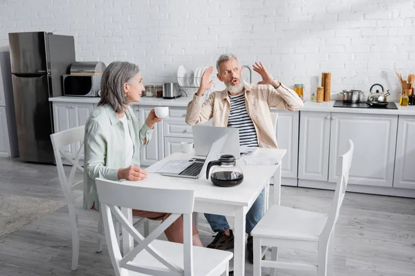 Зрілий чоловік розмовляє з дружиною з кавою біля паперу і ноутбуків на кухні — стокове фото