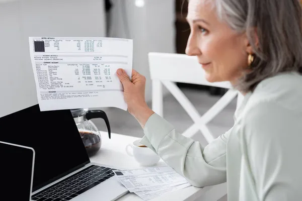 Side view of blurred mature woman holding bills near laptops at home — Stock Photo