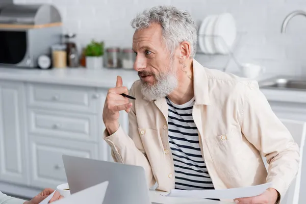 Homme mûr avec du papier gesticulant près de la femme et des ordinateurs portables à la maison — Photo de stock