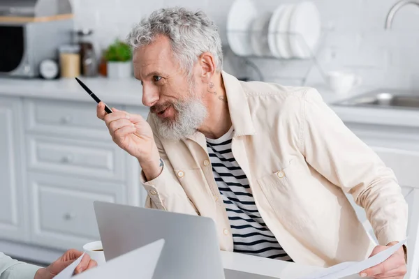 Homme souriant tenant des documents et parlant à sa femme près du café et des ordinateurs portables dans la cuisine — Photo de stock
