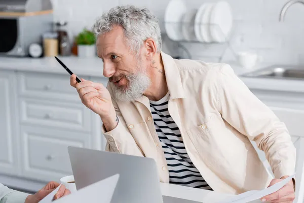 Sonriente hombre sosteniendo documentos y hablando con su esposa cerca de café y computadoras portátiles en la cocina - foto de stock