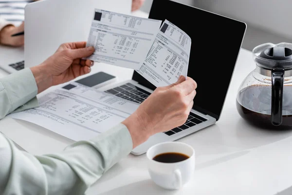 Vista recortada de la mujer sosteniendo facturas cerca de la computadora portátil con pantalla en blanco y taza de café en casa - foto de stock