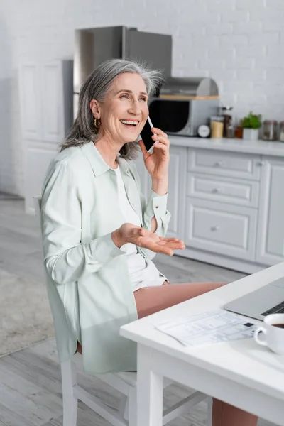 Sorridente donna matura che parla su smartphone vicino laptop, caffè sfocato e bollette in cucina — Foto stock