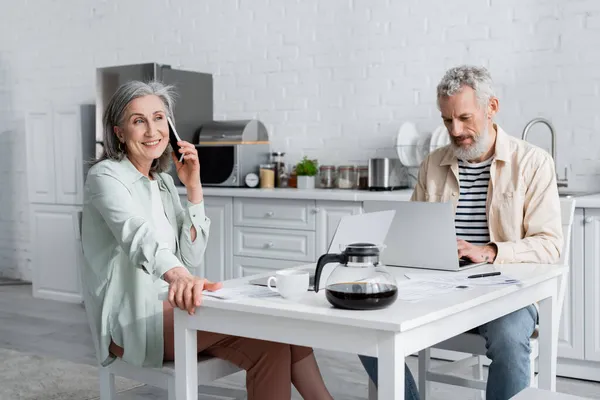 Donna matura che parla sullo smartphone vicino al marito usando laptop e bollette sul tavolo in cucina — Foto stock