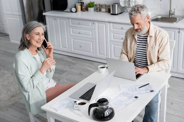 Высокоугольный вид мужчины, использующего ноутбук рядом с кофе, счетами и женой, говорящей по смартфону на кухне — стоковое фото
