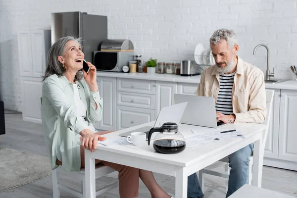 Mulher madura falando no celular perto do marido usando laptop, contas e cafeteira na mesa — Fotografia de Stock