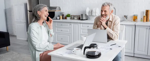 Uomo sorridente guardando la moglie allegra che parla sul cellulare vicino a laptop, caffè e bollette in cucina, banner — Foto stock