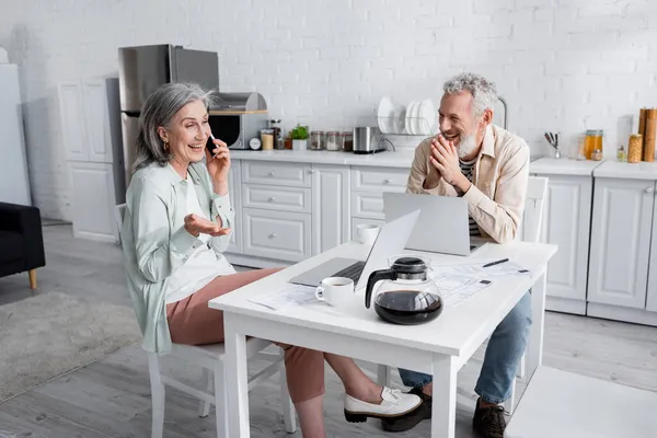 Mujer madura hablando en smartphone cerca de marido alegre, ordenadores portátiles y facturas en la cocina - foto de stock