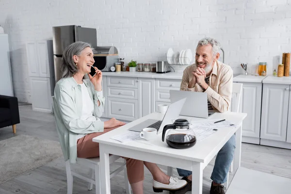 Позитивная зрелая женщина разговаривает на смартфоне и смотрит на мужа возле ноутбуков и счетов на кухне — стоковое фото