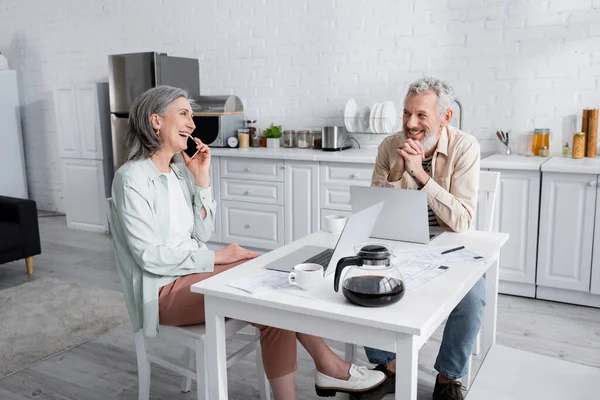 Positivo donna matura parlando su smartphone e guardando il marito vicino a computer portatili e bollette in cucina — Foto stock