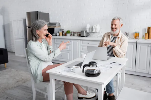 Lächelnde reife Frau greift zum Handy und zeigt in der Nähe von Laptops und Rechnungen in der Küche auf ihren Mann — Stockfoto