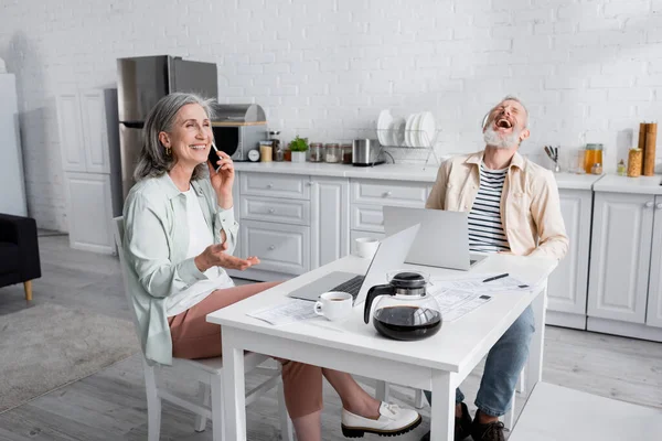 Lächelnde reife Frau spricht auf Smartphone neben fröhlichem Ehemann, Laptops und Rechnungen zu Hause — Stockfoto