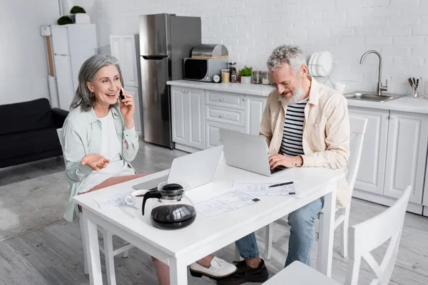 Mature man using laptop near wife talking on smartphone, coffee and bills in kitchen — Stock Photo