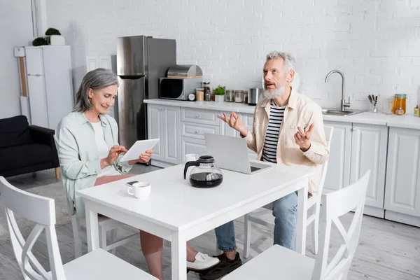 Pensativo hombre maduro sentado cerca de la computadora portátil, café y esposa con tableta digital en la cocina - foto de stock