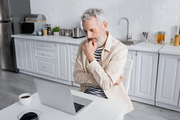 Pensive freelancer looking at laptop near cup of coffee in kitchen — Stock Photo