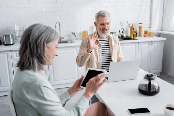 Homme souriant ayant appel vidéo sur ordinateur portable près de café et femme avec tablette numérique à la maison — Photo de stock