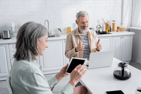 Mature homme montrant pouces levés lors d'un appel vidéo sur ordinateur portable près de café et femme avec tablette numérique à la maison — Photo de stock