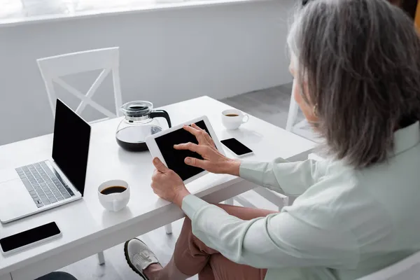Размытая зрелая женщина с помощью цифрового планшета возле устройств и кофе на кухне — стоковое фото